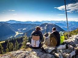 $PageTeaserImageTitle Familie sitzt auf einem Berggipfel mit Blick auf Tannenwälder und Gipfel