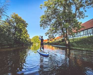 Ein Hotelgast fährt im Wassersporturlaub mit einem Stand Up Board auf dem Fluss Ein Hotelgast fährt im Wassersporturlaub mit einem Stand Up Board auf dem Fluss