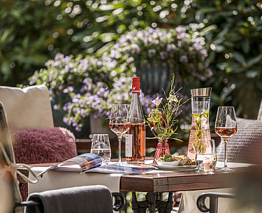 Tisch mit Weinglas, Weinkaraffe, Flasche und Snacks im Garten mit Blumen im Hintergrund
