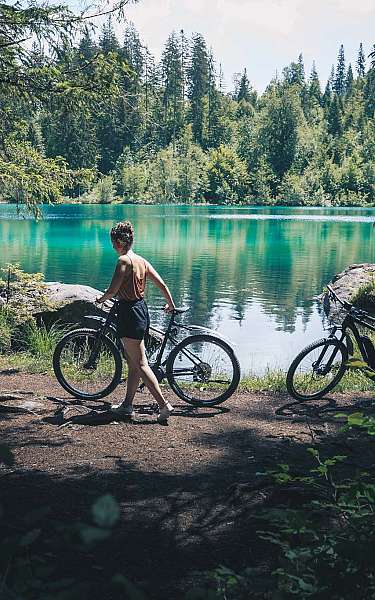 Frau mit zwei Fahrrädern an einem Waldsee mit grünen Bäumen im Hintergrund